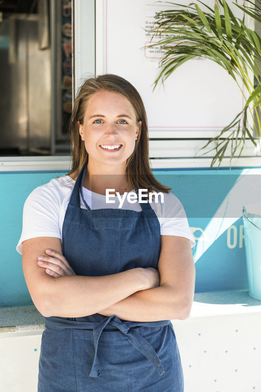 Portrait of smiling young female owner standing with arms crossed against food truck