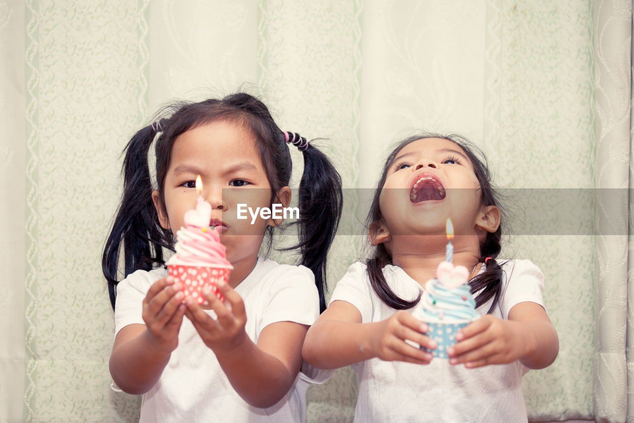 Sisters holding cupcakes against curtain during birthday