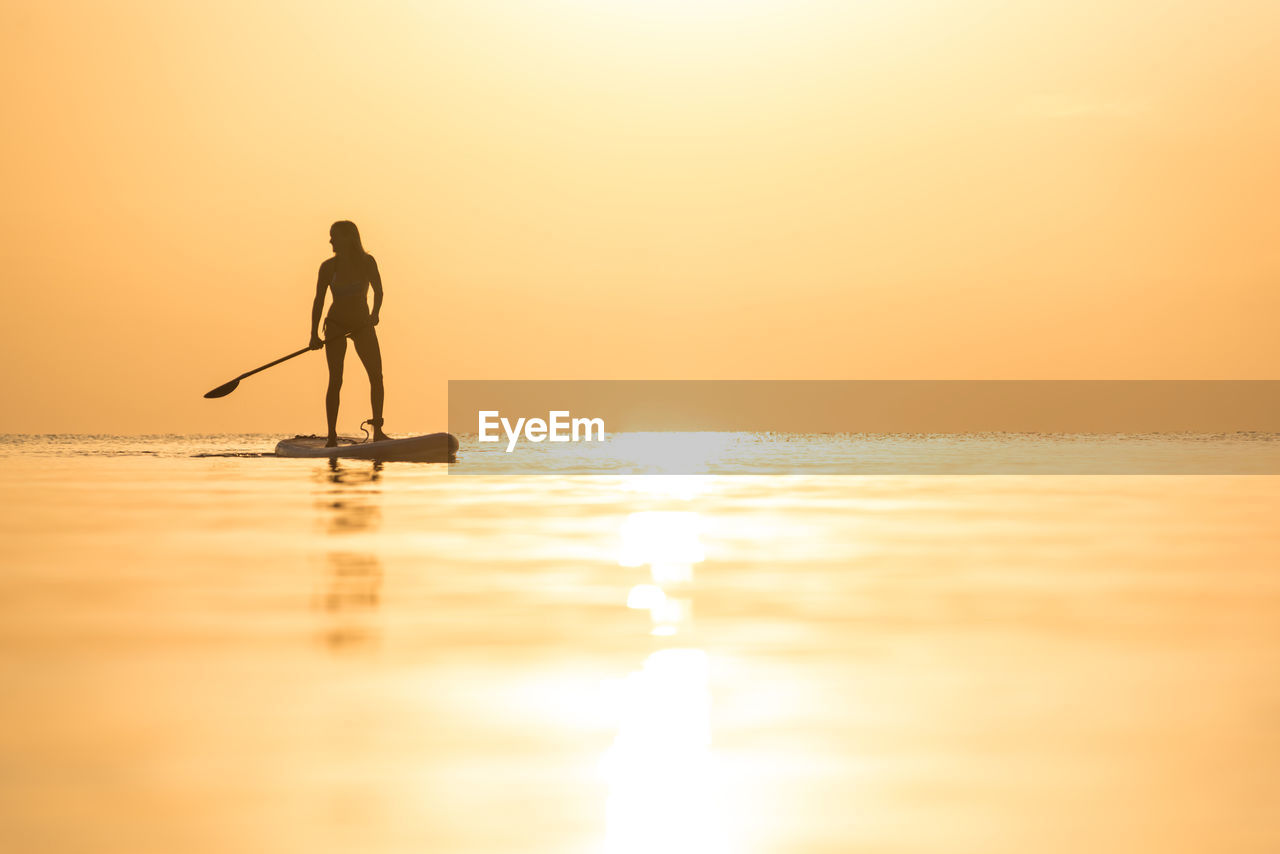 Silhouette of unrecognizable female surfer standing on paddleboard and rowing against spectacular sun in sunset sky