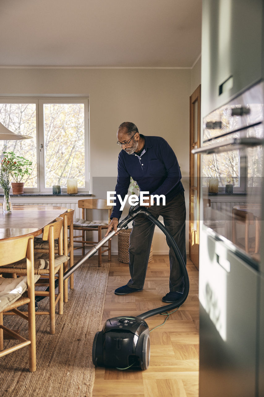 Senior man cleaning carpet with vacuum cleaner while standing at home