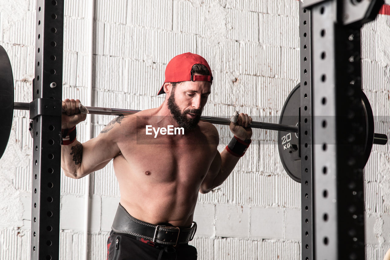 Confident shirtless sportsman standing with heavy barbell and preparing for doing squats during workout in gym
