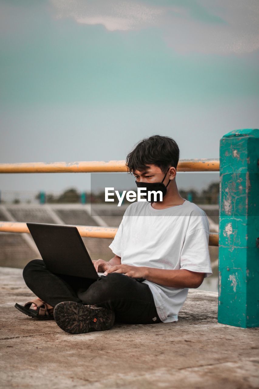 Young man using laptop wearing flu mask sitting by railing outdoors against sky