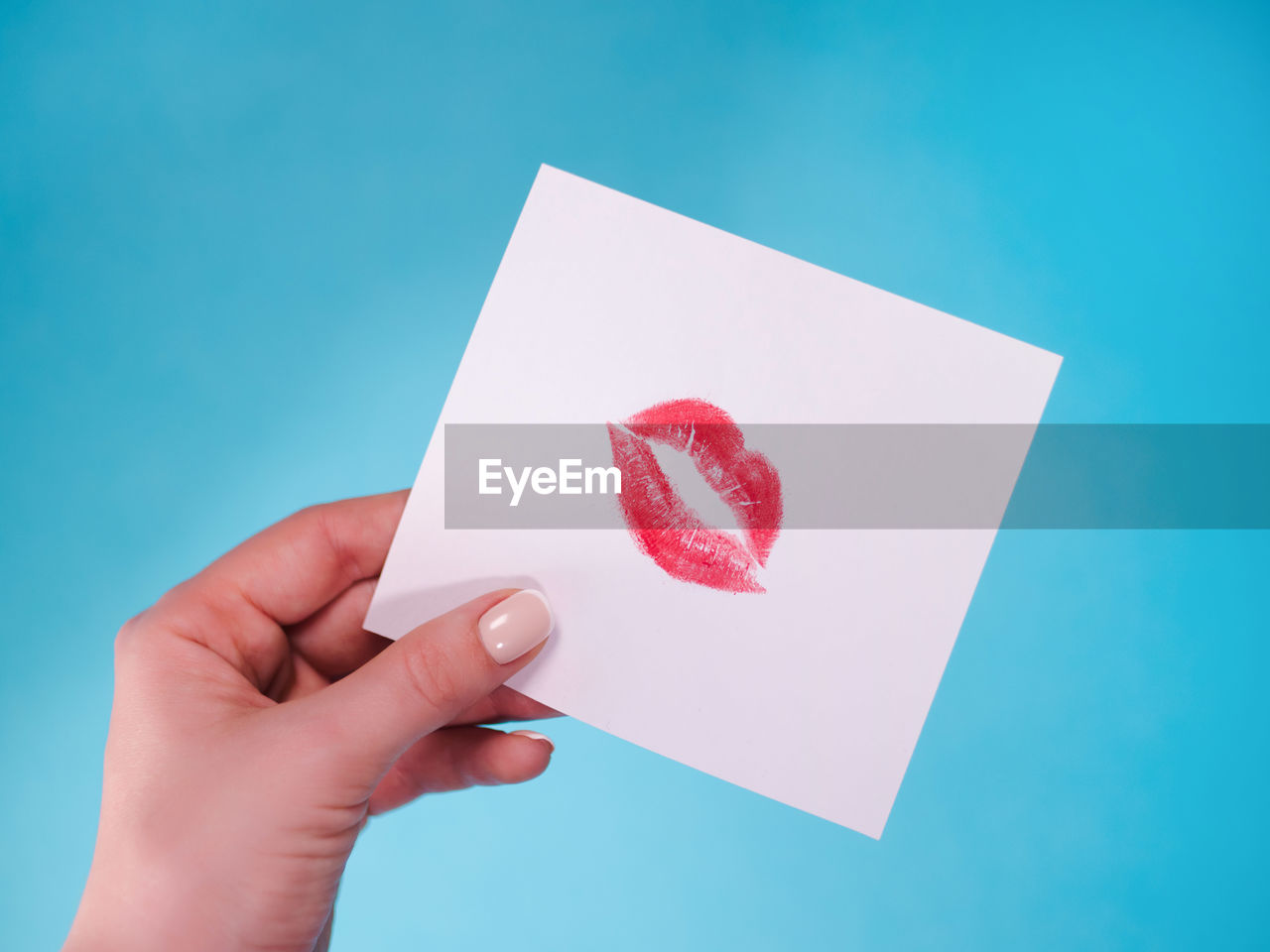 Cropped hand of woman holding paper with lipstick kiss against blue background