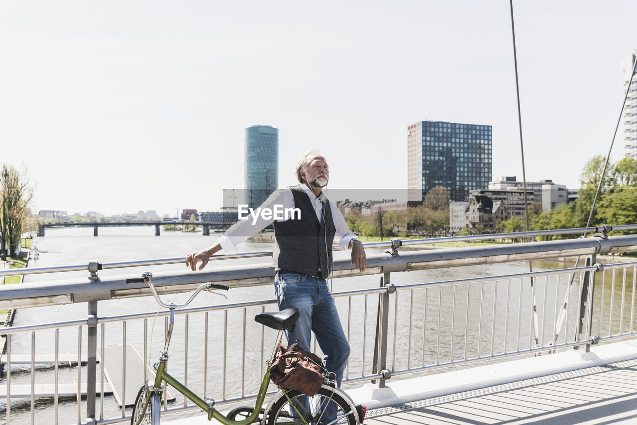 Mature man with bicycle listening to music on bridge in the city