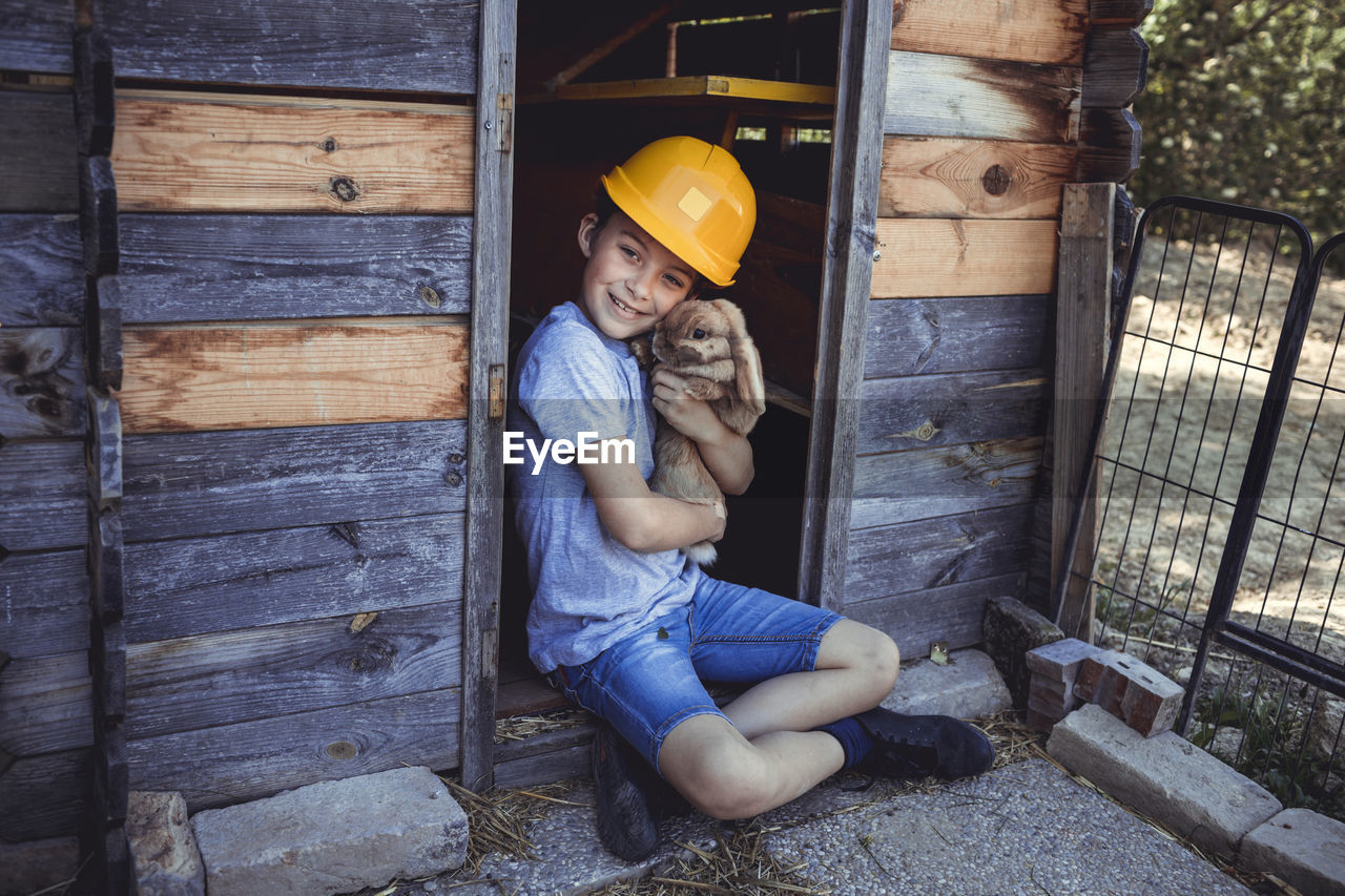 Cute boy in yellow hardhat holding rabbit in wooden hutch