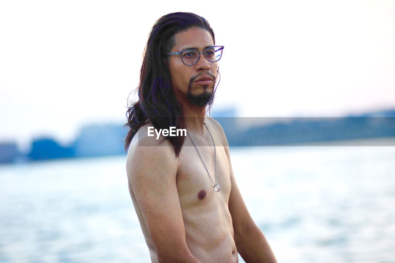 Shirtless man looking away while standing against lake