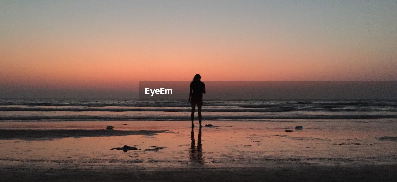 Silhouette woman standing at beach against orange sky