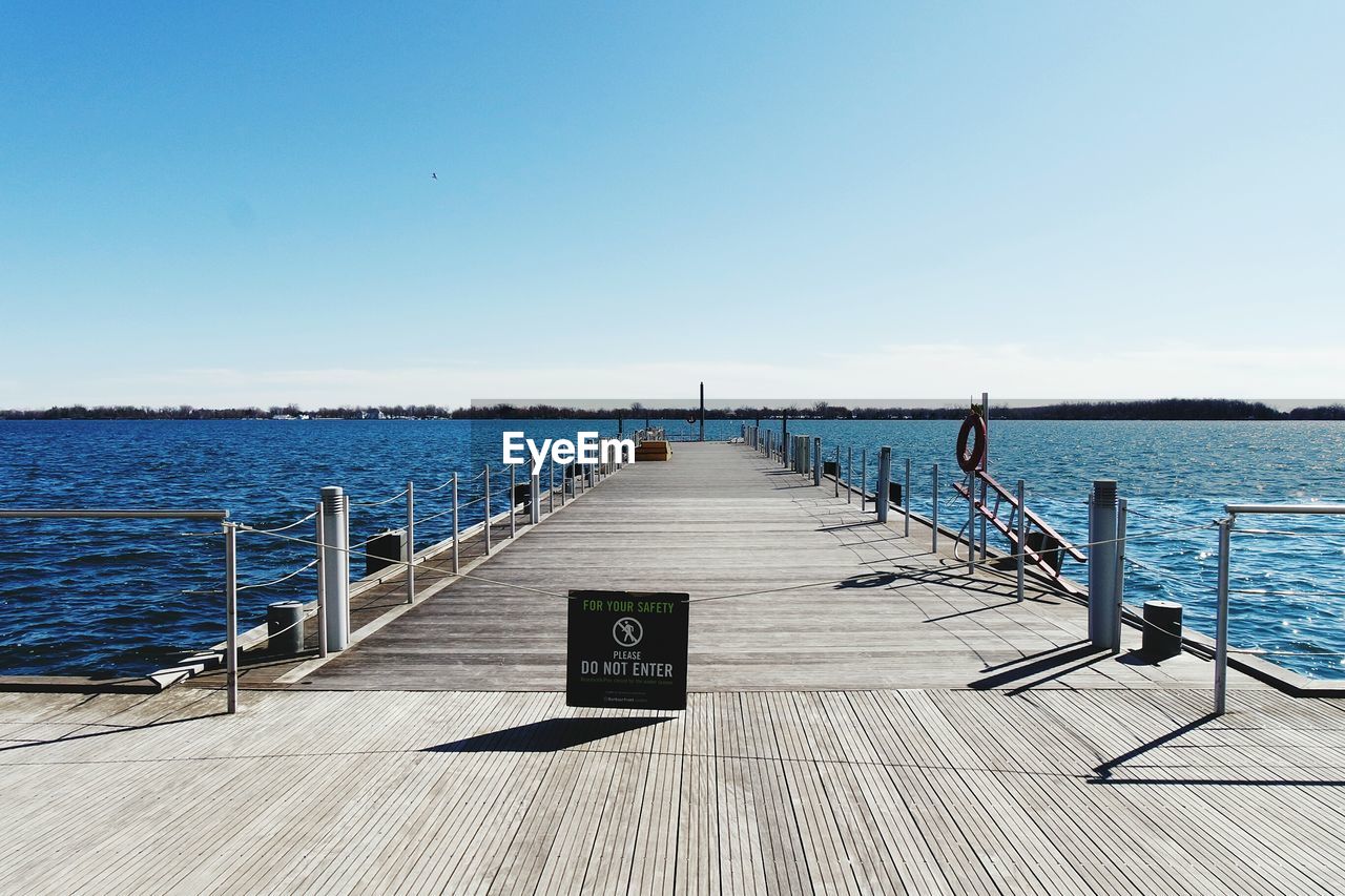 Warning sign on pier amidst river against clear sky on sunny day