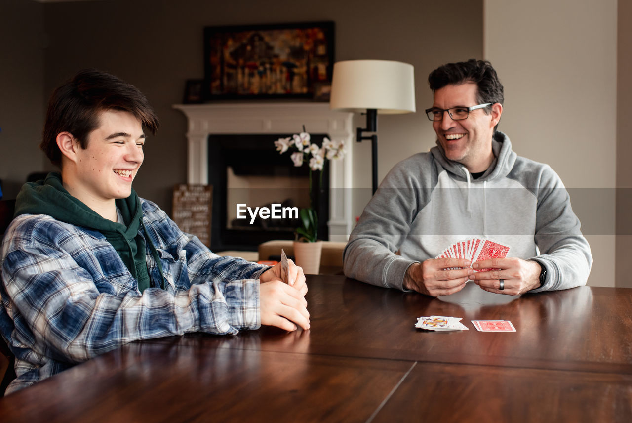 Father and teenage son laughing as they play cards at the table.