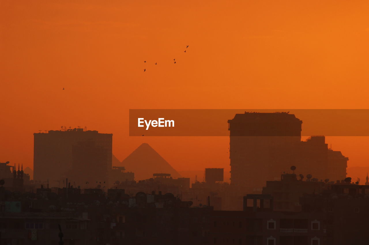 Pyramids and buildings in city against orange sky in egypt