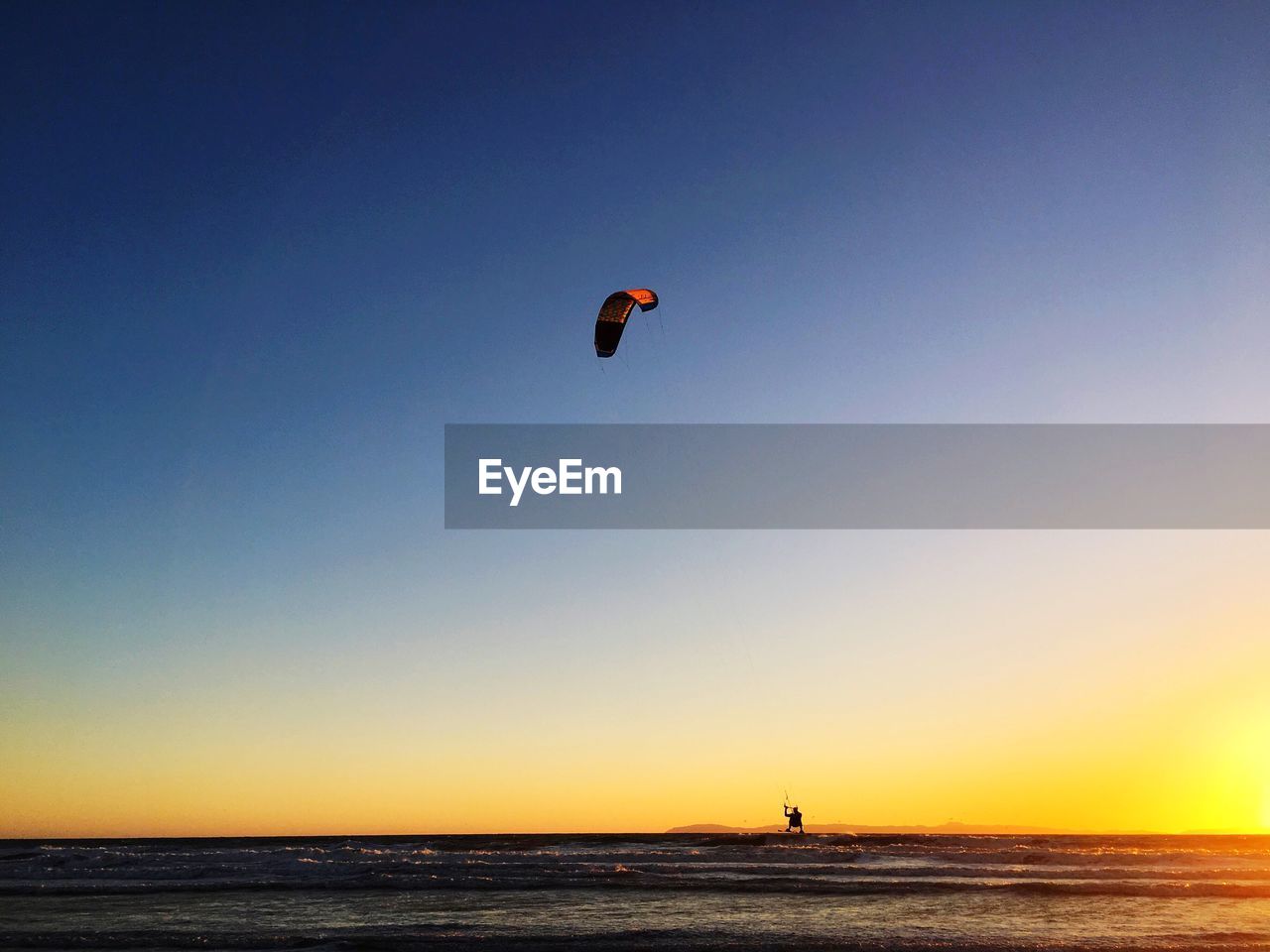 PEOPLE PARAGLIDING AT BEACH AGAINST SKY DURING SUNSET