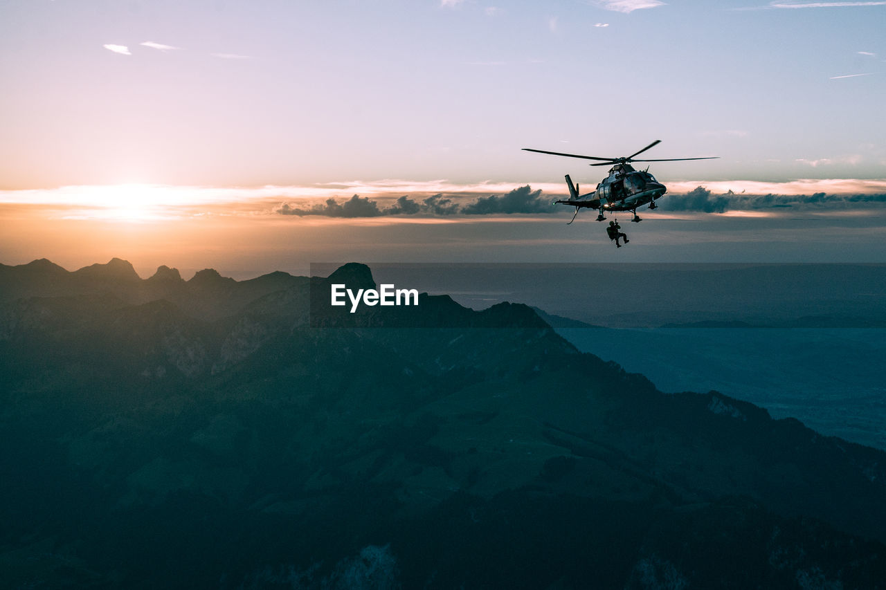 Helicopter flying over mountains against sky at sunset