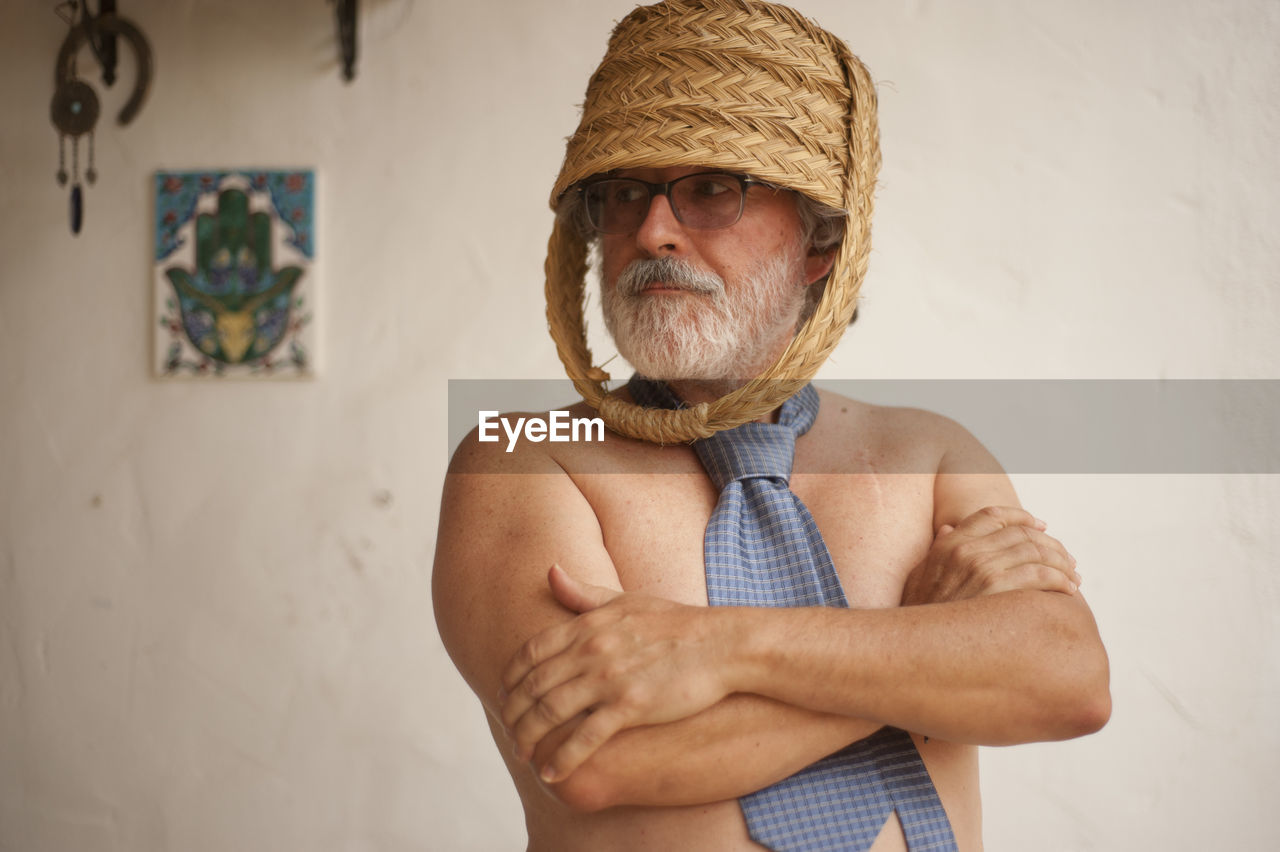 Shirtless senior man with basket on head standing against wall at home