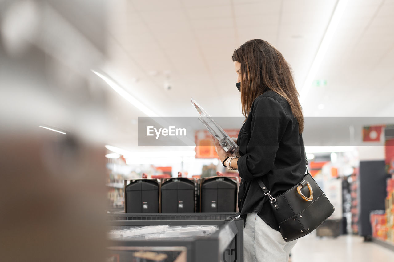 Side view of female customer in black protective mask and gloves choosing products in supermarket during coronavirus pandemic