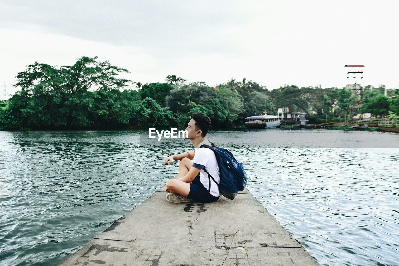 Young man sitting on jetty over lake against sky