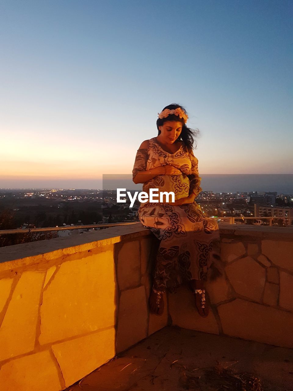 Full length of pregnant woman sitting on retaining wall against sky