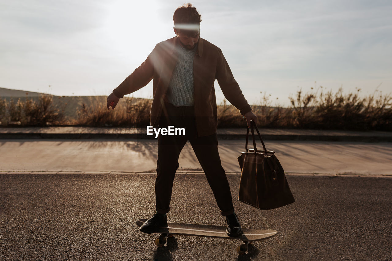 Full body serious young bearded male skater in trendy clothes with leather bag riding skateboard along asphalt road