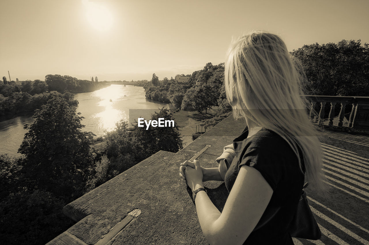 Woman standing at railing by river against sky during sunset