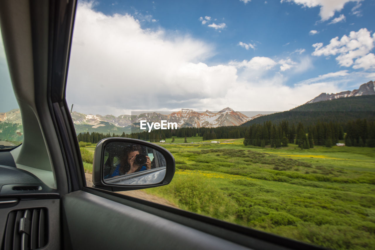 Reflection of woman photographing in side-view mirror against mountains