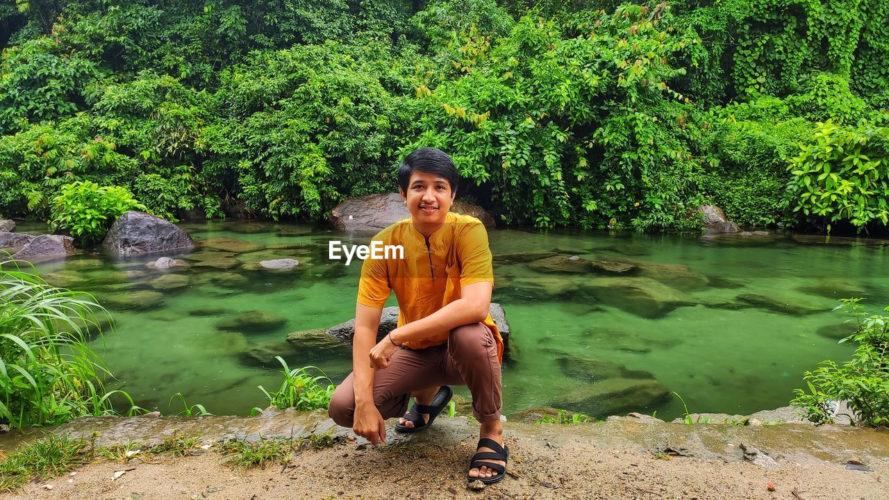 Portrait of man crouching by lake against trees