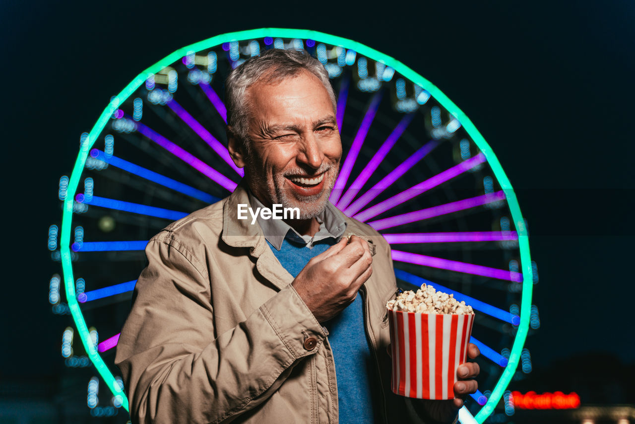 Portrait of senior man eating popcorn while standing against ferris wheel at night