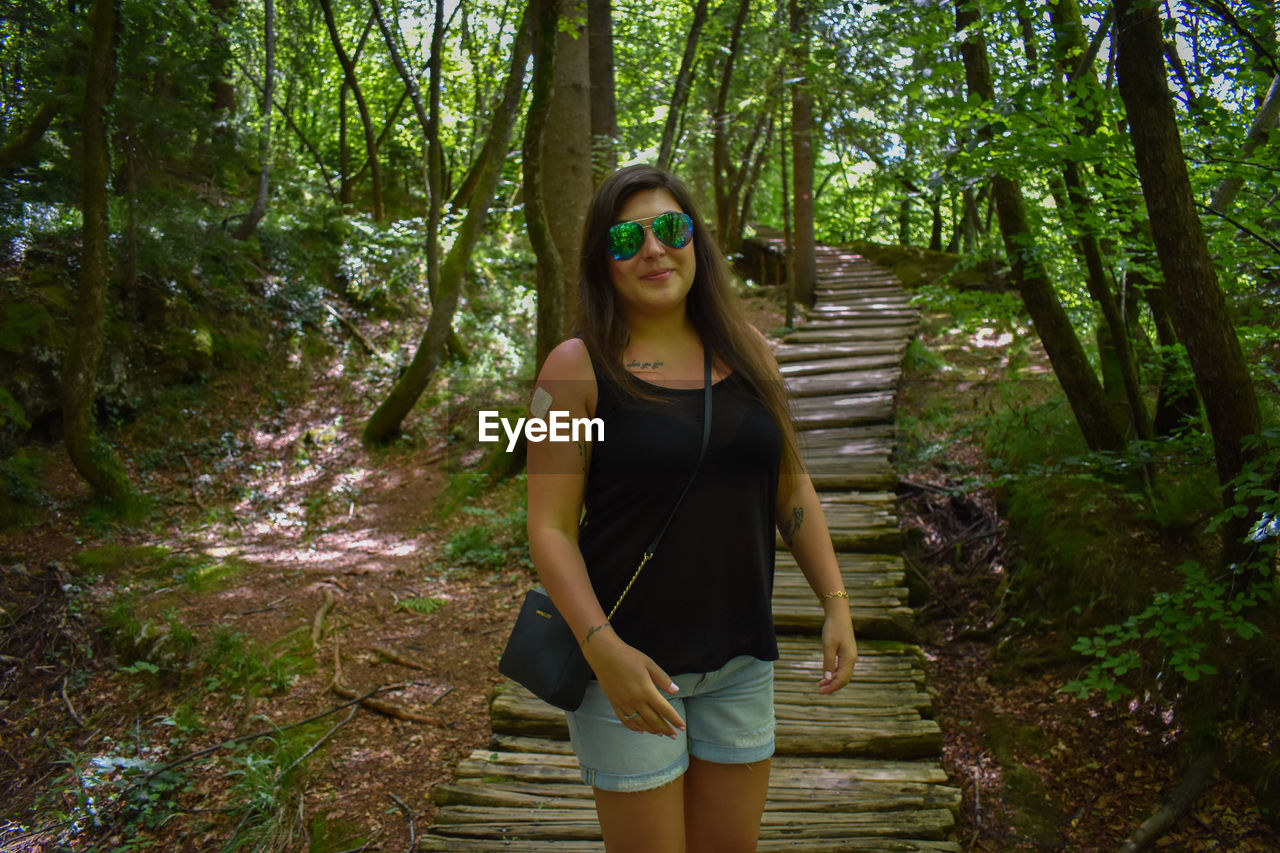 Portrait of smiling mid adult woman wearing sunglasses while standing on boardwalk amidst trees in forest