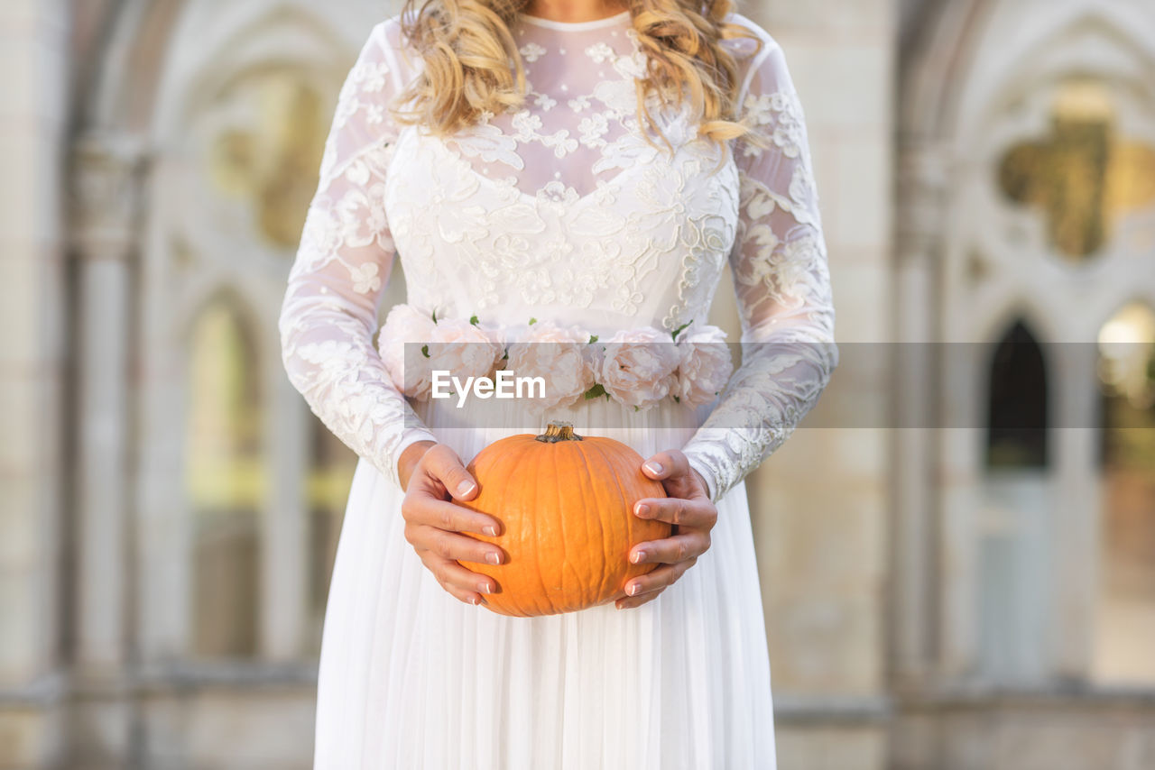 MIDSECTION OF WOMAN HOLDING PUMPKIN WHILE STANDING AGAINST ORANGE BACKGROUND