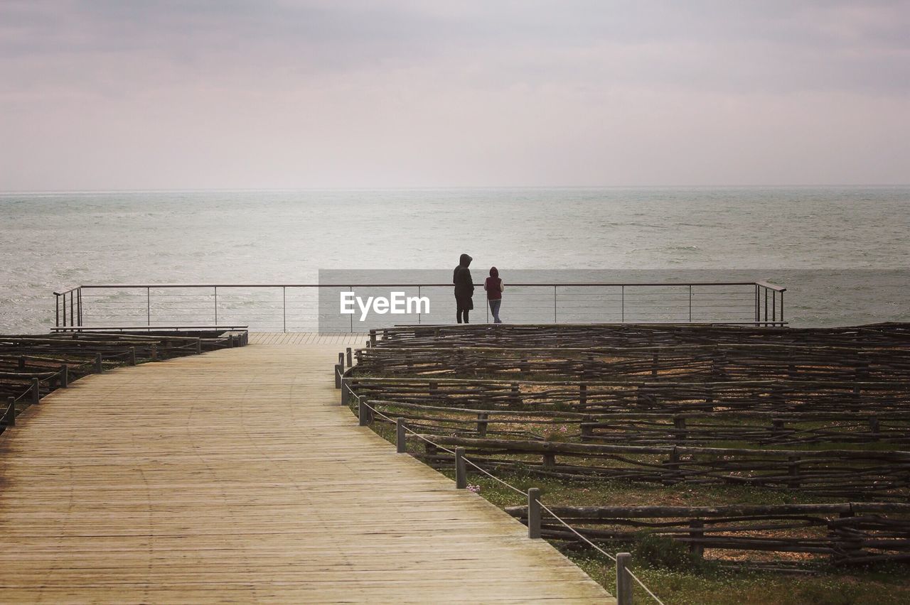 Rear view of mother with son standing at beach against cloudy sky