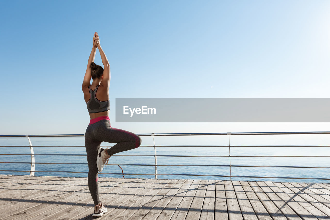 Rear view of woman exercising by railing while standing against sea