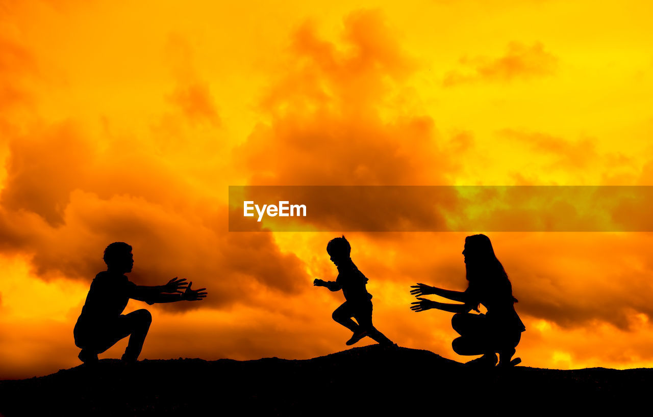 Silhouette parents playing with son on mountain against orange cloudy sky during sunset