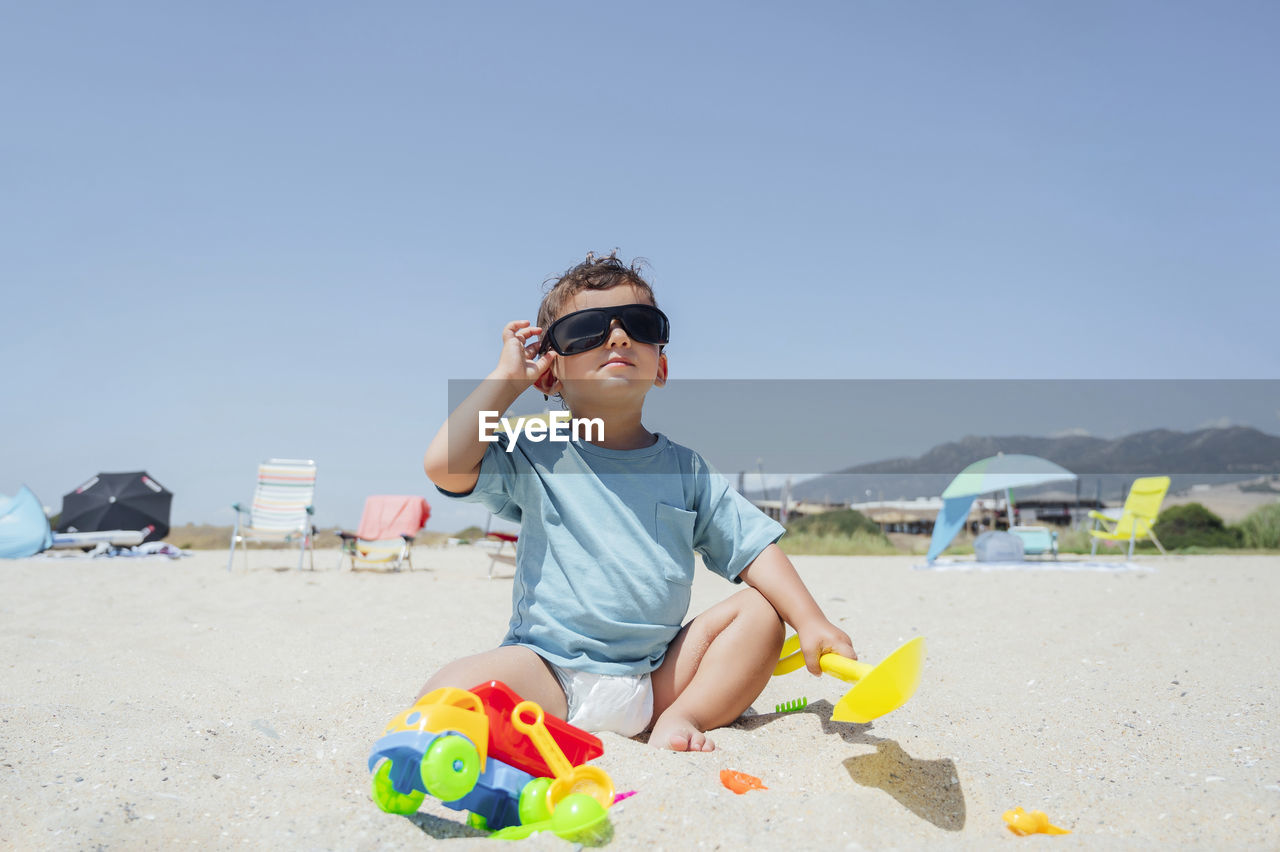 Boy adjusting sunglasses sitting with toys on sand at beach