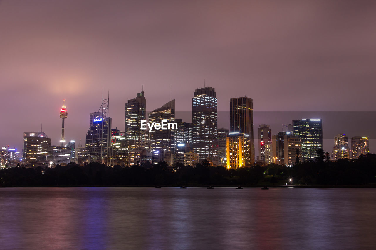 Illuminated buildings by river against sky at night