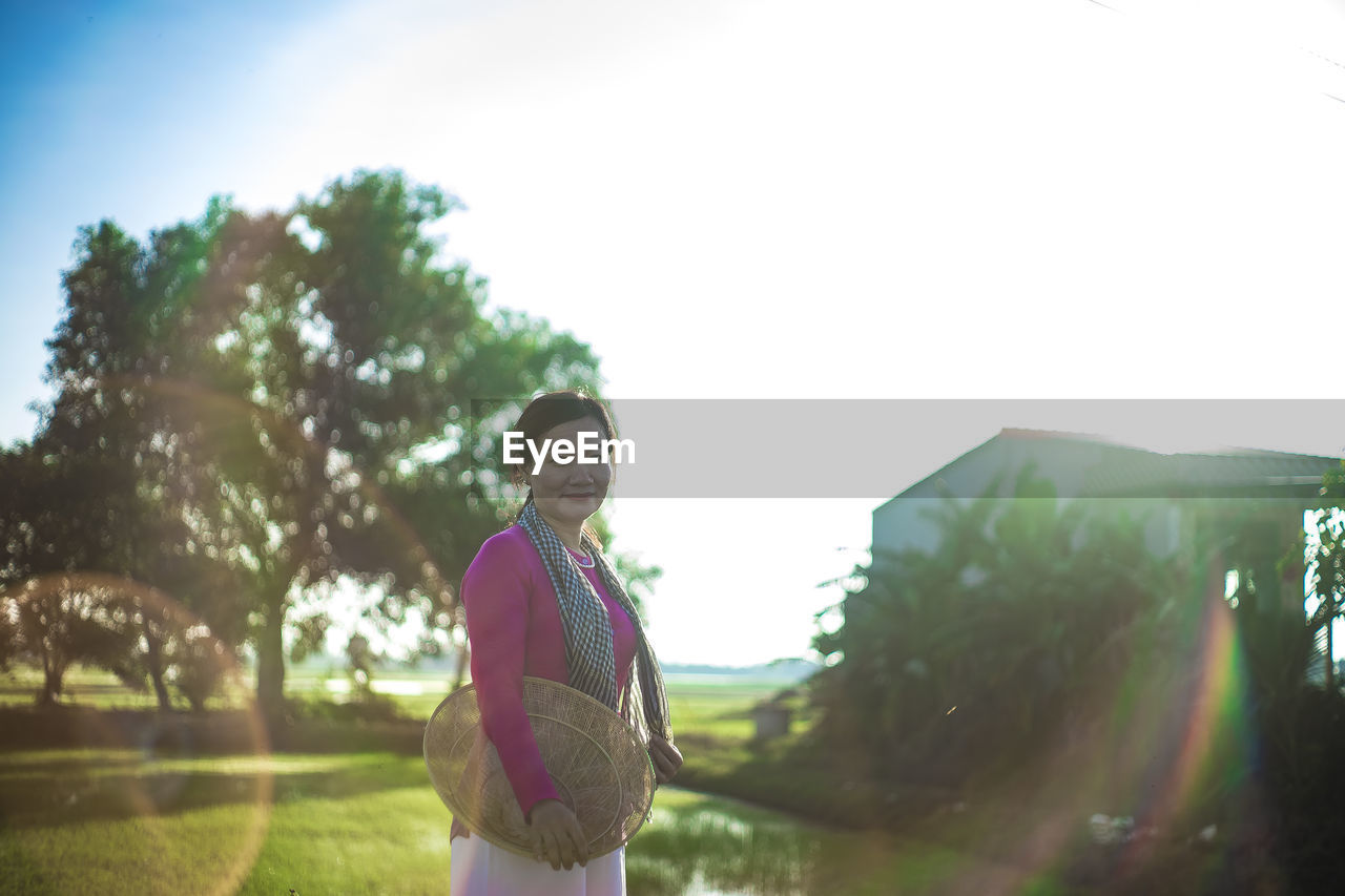 one person, nature, lens flare, sunlight, sky, adult, plant, day, copy space, leisure activity, standing, women, back lit, lifestyles, tree, young adult, clothing, sports, outdoors, person, grass, casual clothing, smiling, sunbeam, happiness, looking, summer, sunny, female, three quarter length, waist up, relaxation, landscape, emotion