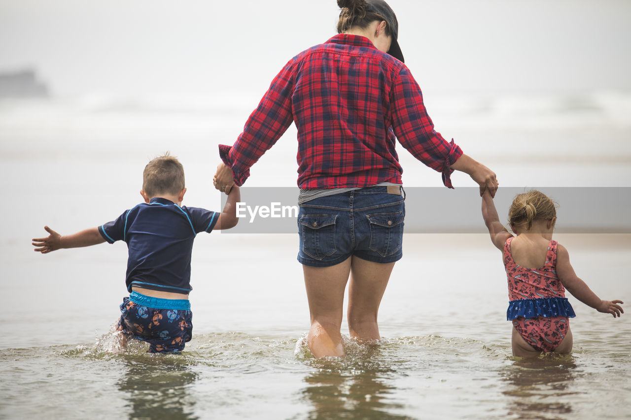 Rear view of mother helping kids wade through water at the beach.