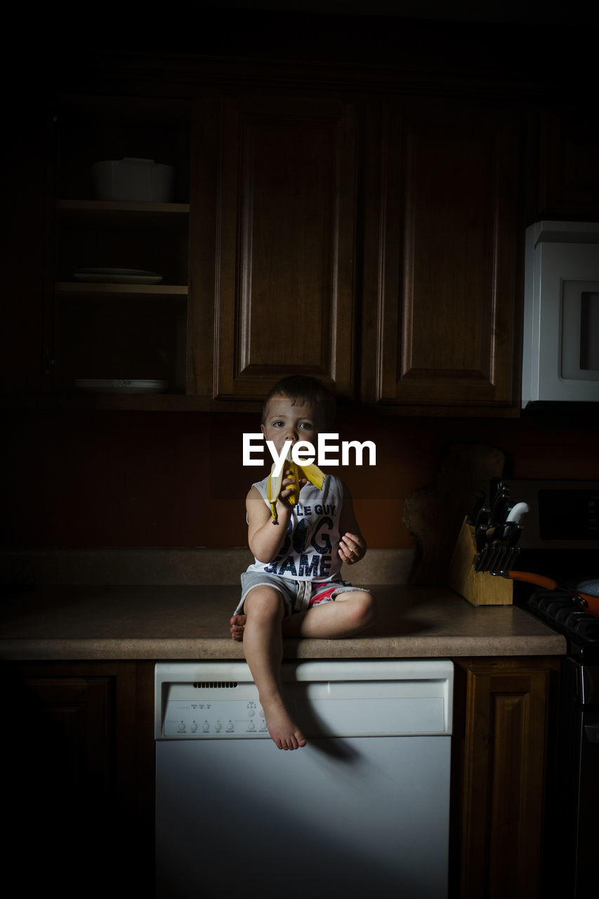 Portrait of boy eating banana while sitting on kitchen counter at home