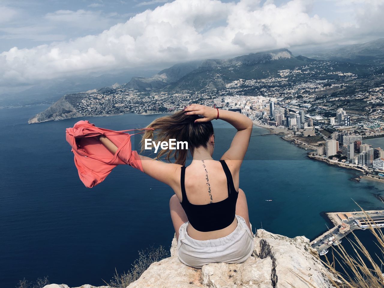 Rear view of woman looking at cityscape sitting at cliff against sky