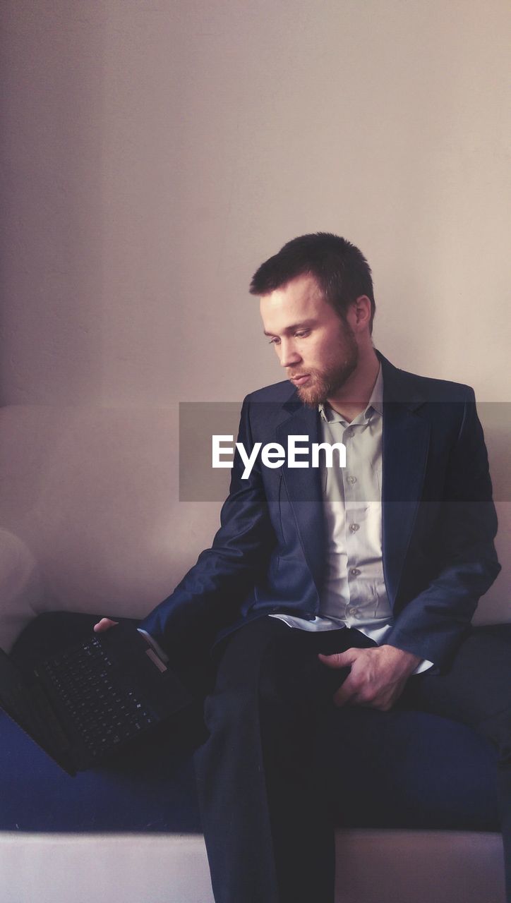 Businessman using laptop while sitting on sofa against wall in office