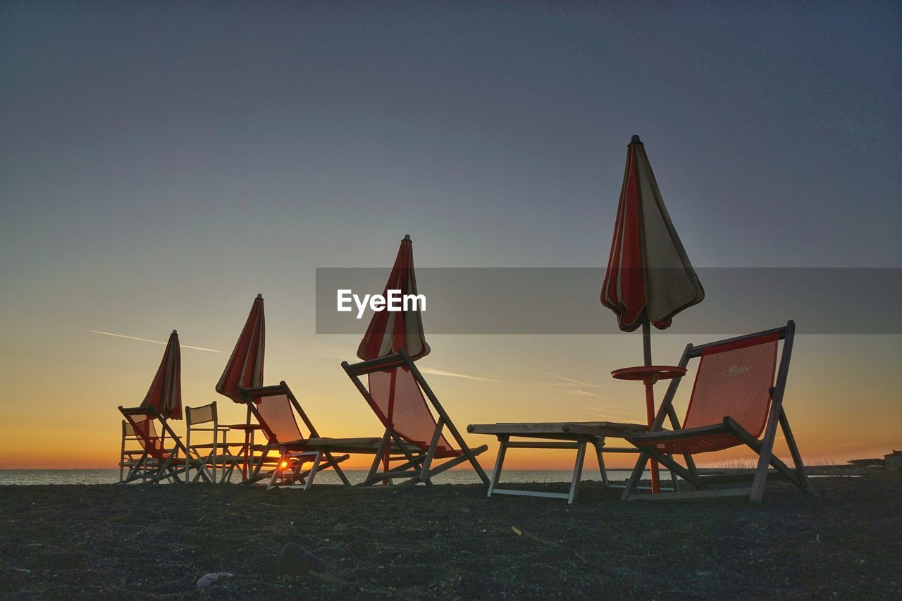 Closed parasols and chairs at beach against clear sky during sunset