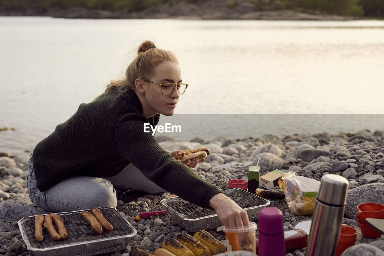 Young woman preparing food while sitting at beach during sunset