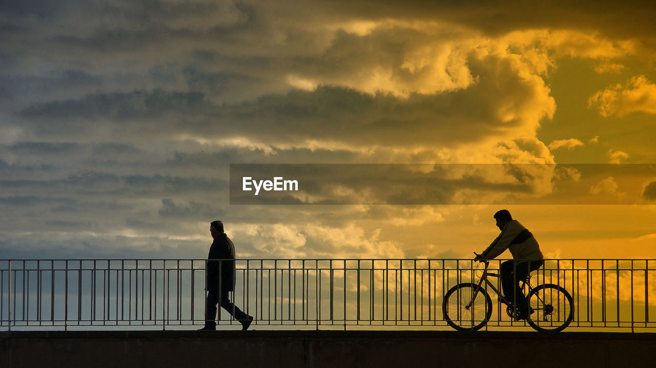 Man riding bicycle on bridge against cloudy sky during sunset