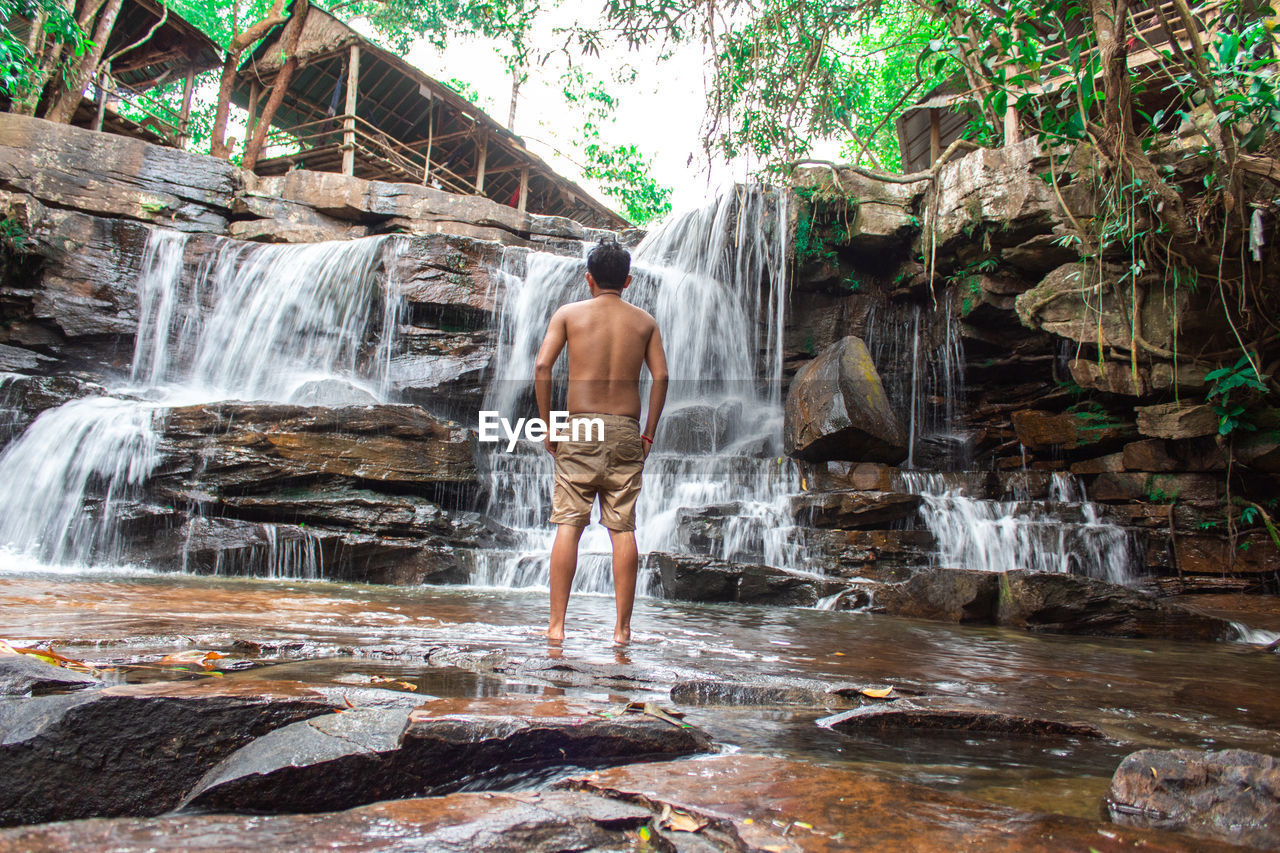Rear view of shirtless man standing by waterfall in forest