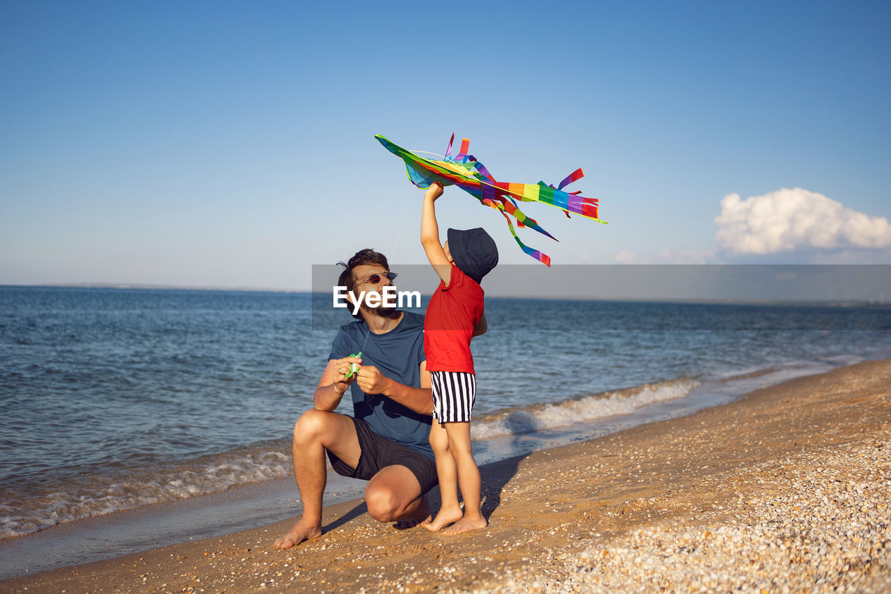 Father and son are standing on a sandy beach by the sea and launch a toy striped kite in the summer