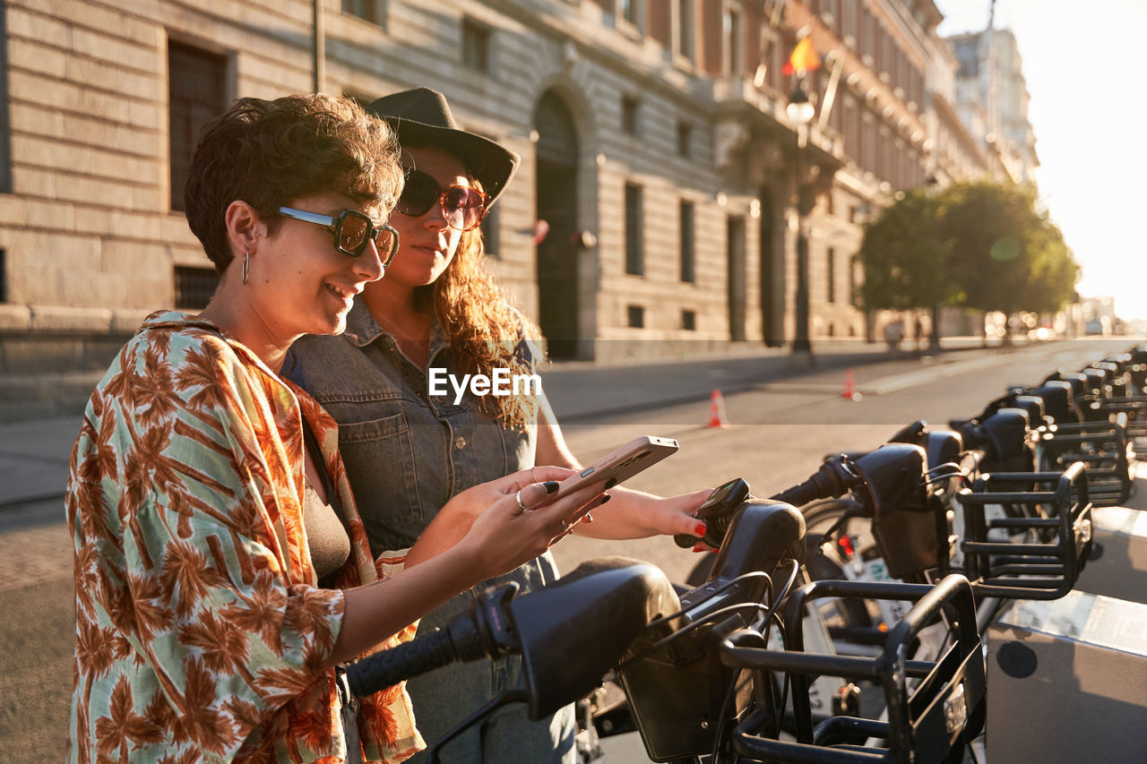 Happy young girlfriends using smartphone to rent electric scooter on sunlit street of madrid at sunset in spain