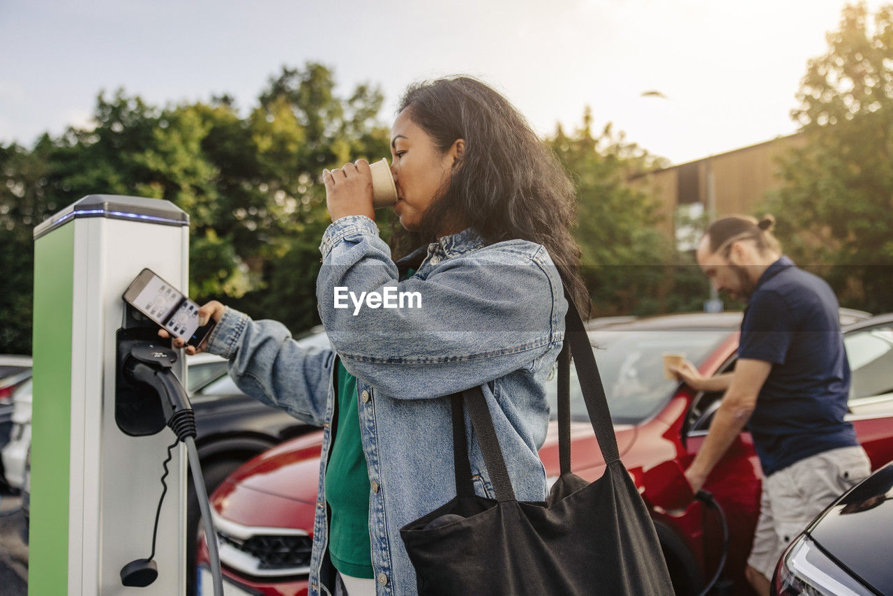 Woman having drink while operating kiosk at charging station