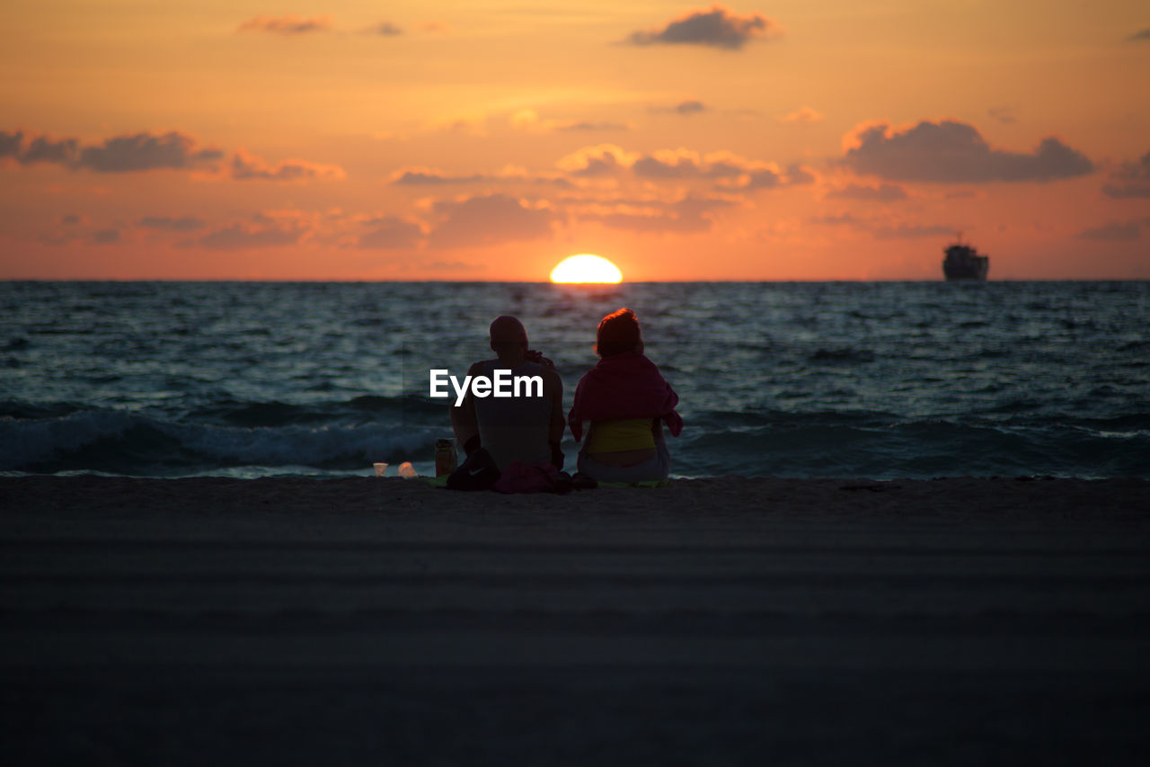 Rear view of men sitting on beach against sky during sunset