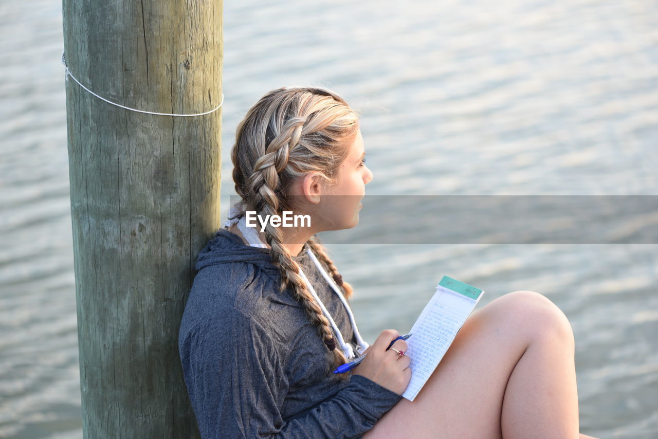 High angle view of thoughtful teenage girl writing on note pad while sitting by lake