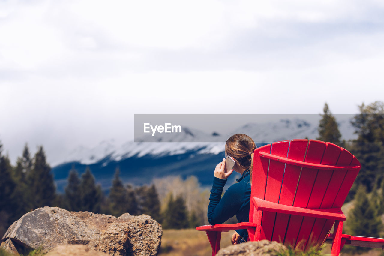 Rear view of woman using smart phone while sitting on chair against snowcapped mountain