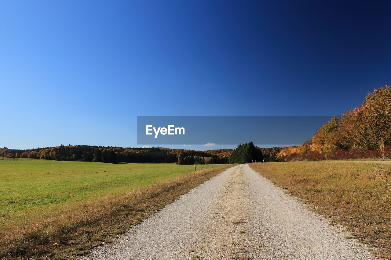 Empty road amidst field against clear blue sky