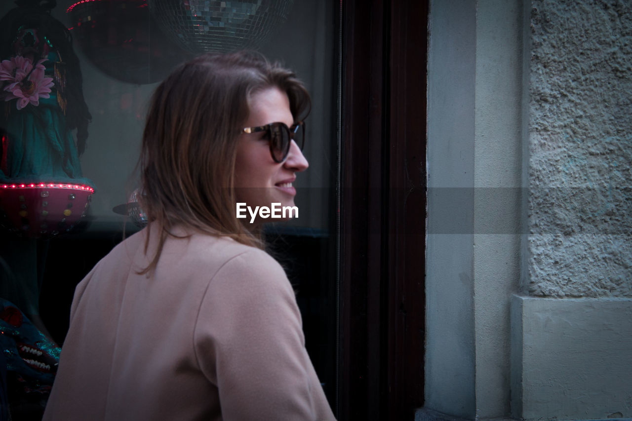 PORTRAIT OF SMILING WOMAN LOOKING AWAY WHILE STANDING BY WINDOW