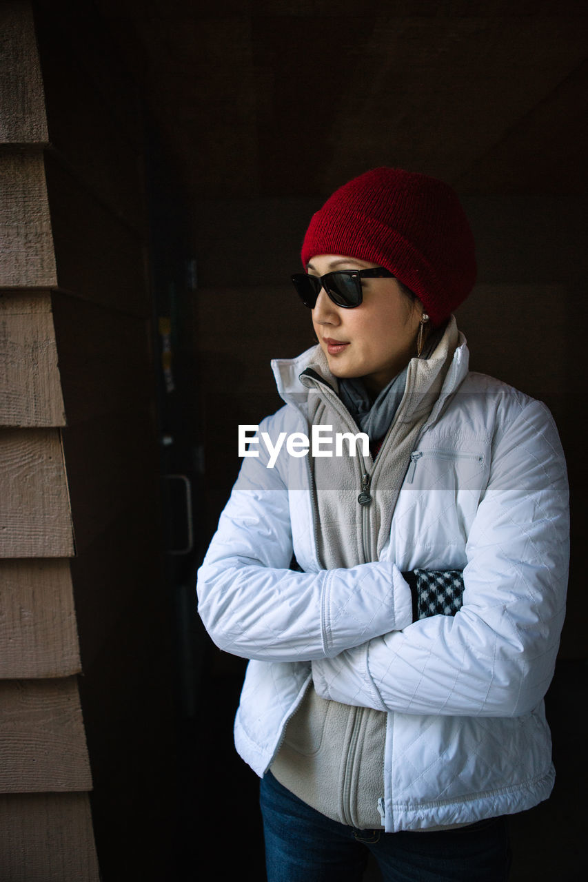 Woman in warm clothing standing with arms crossed by wooden wall